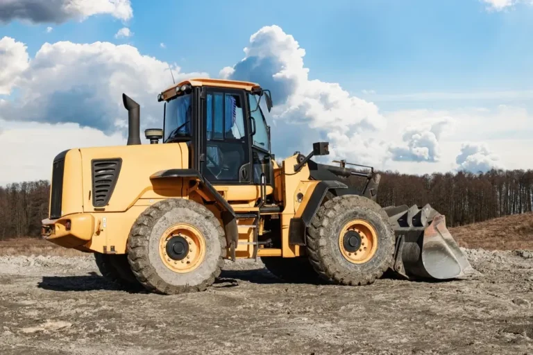 A large yellow front-loader sits idle on a construction site, surrounded by rough terrain under a bright blue sky with fluffy clouds.